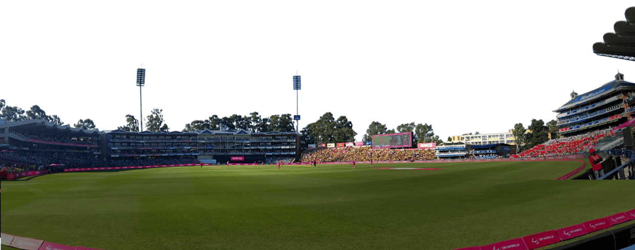 Panoramic view of a cricket stadium with a lush green outfield, stands filled with spectators, and tall floodlights above the field.